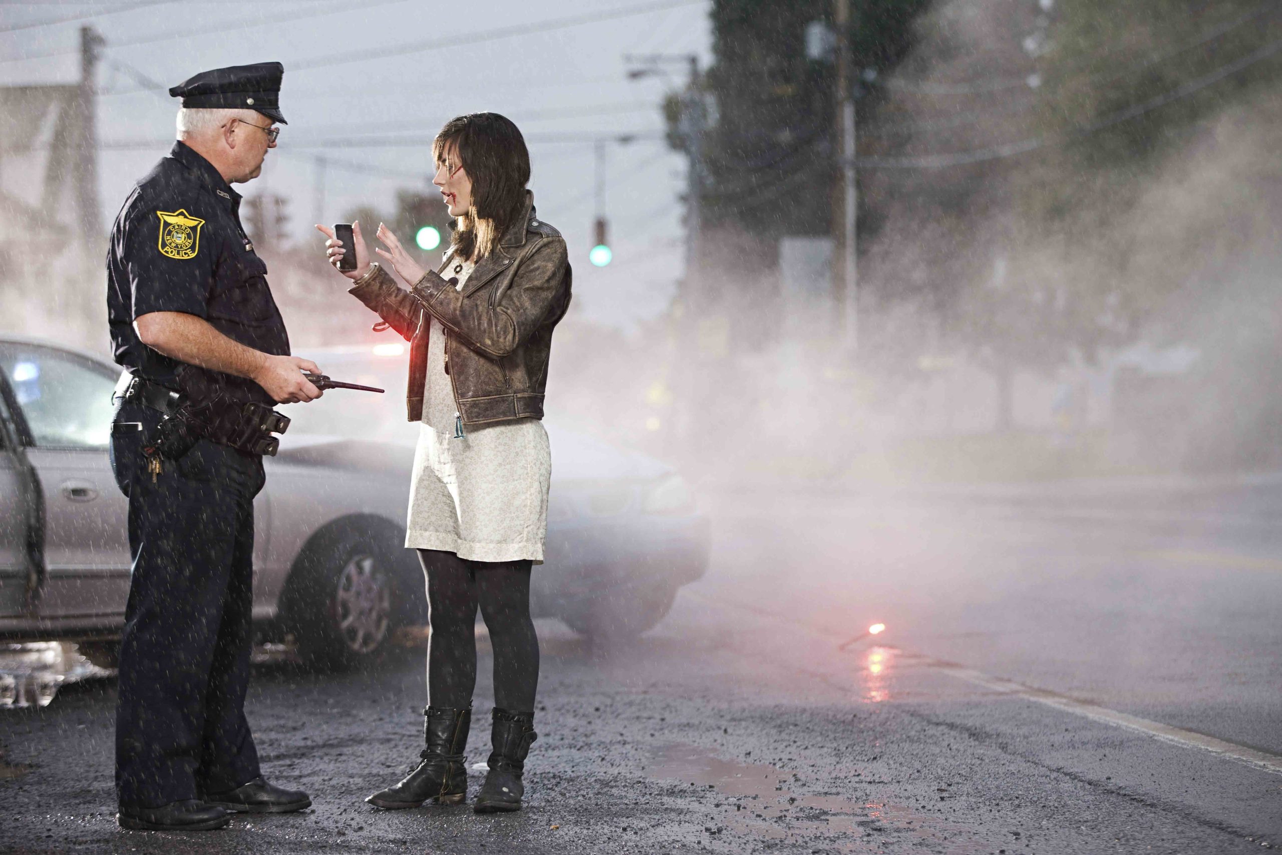 Police officer taking a statement from a woman at a rainy car accident scene with flares and a damaged vehicle