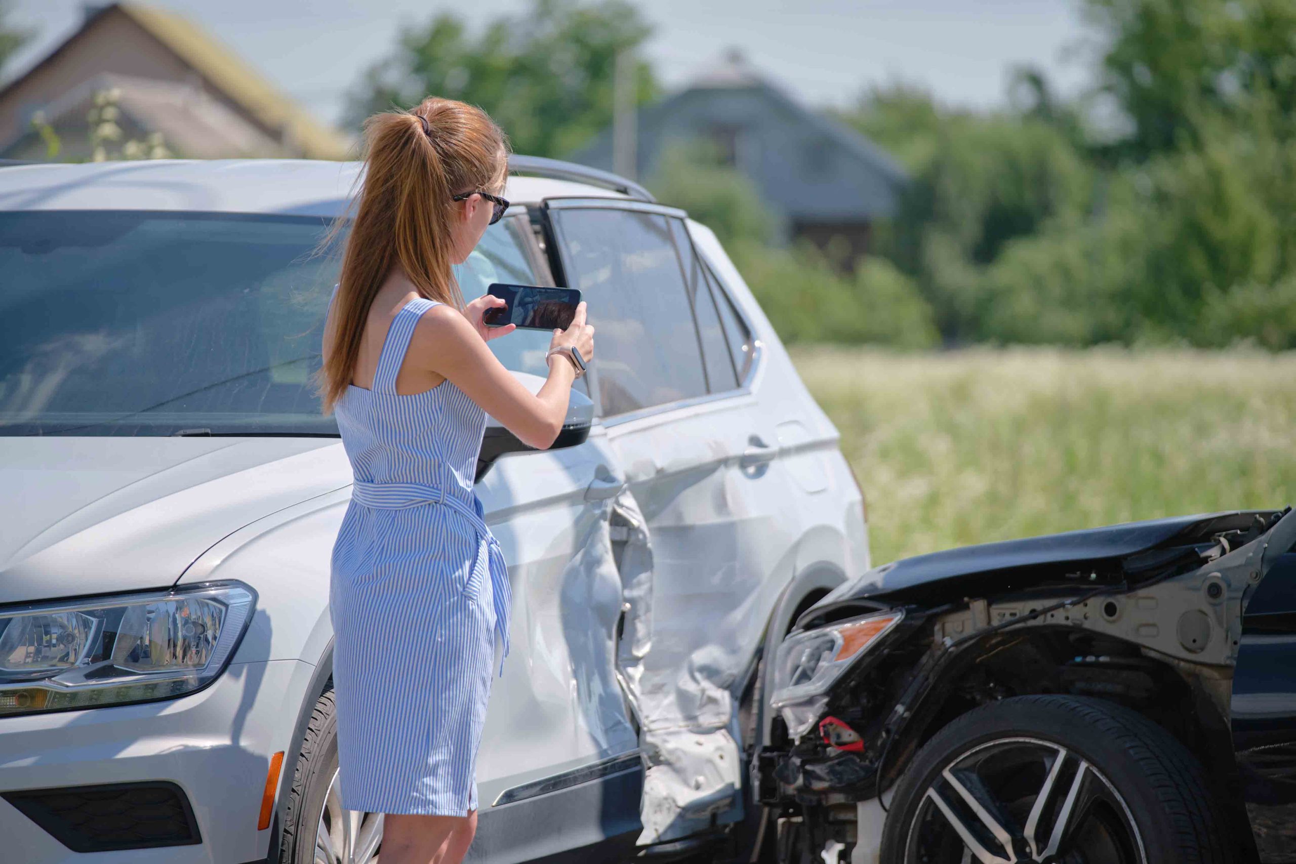 Woman taking photos of car accident damage on her phone, documenting the collision scene. 