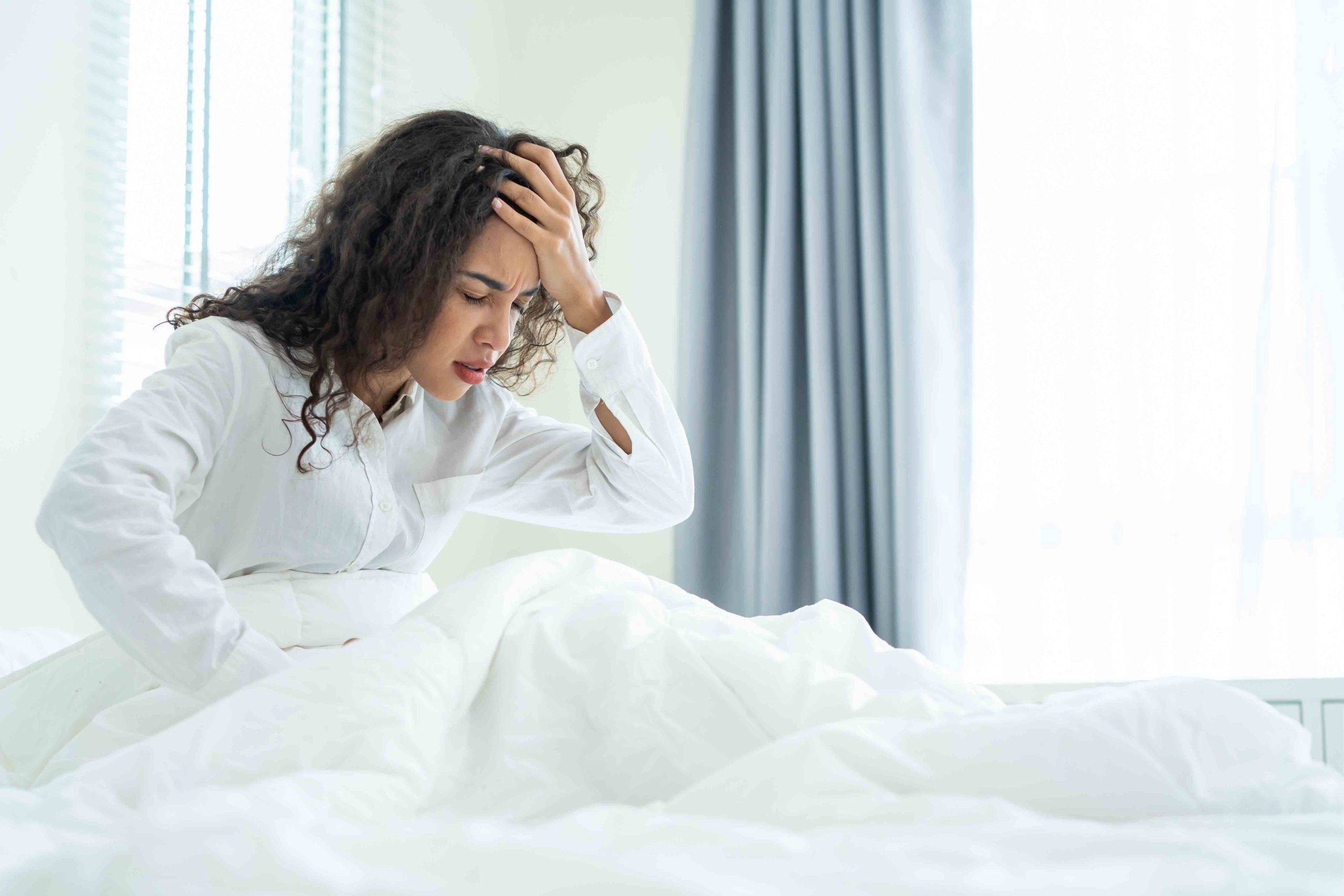 A woman sitting up in bed, holding her head in her hand, indicating pain or discomfort. 