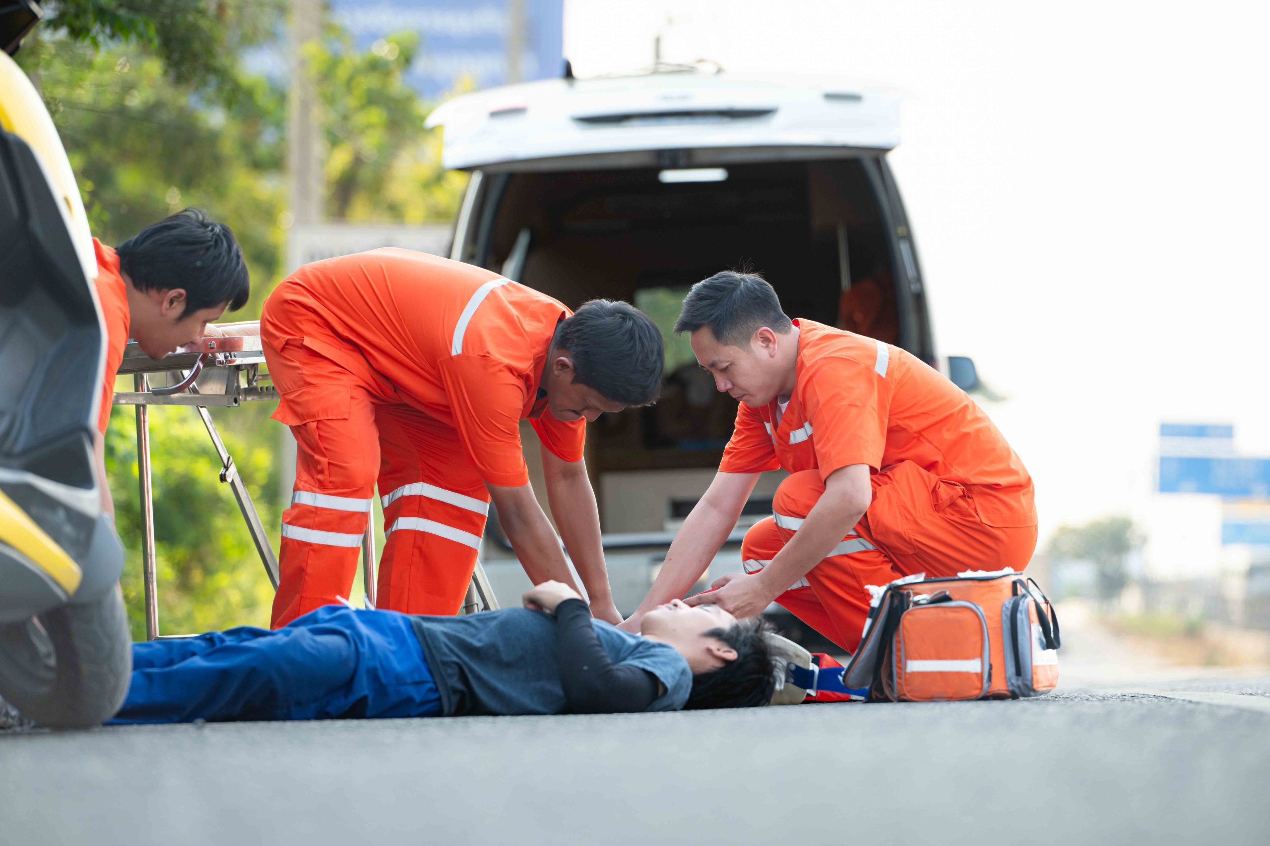 Paramedics in orange uniforms attending to a person lying on the ground next to an open ambulance, providing medical assessment and care. 