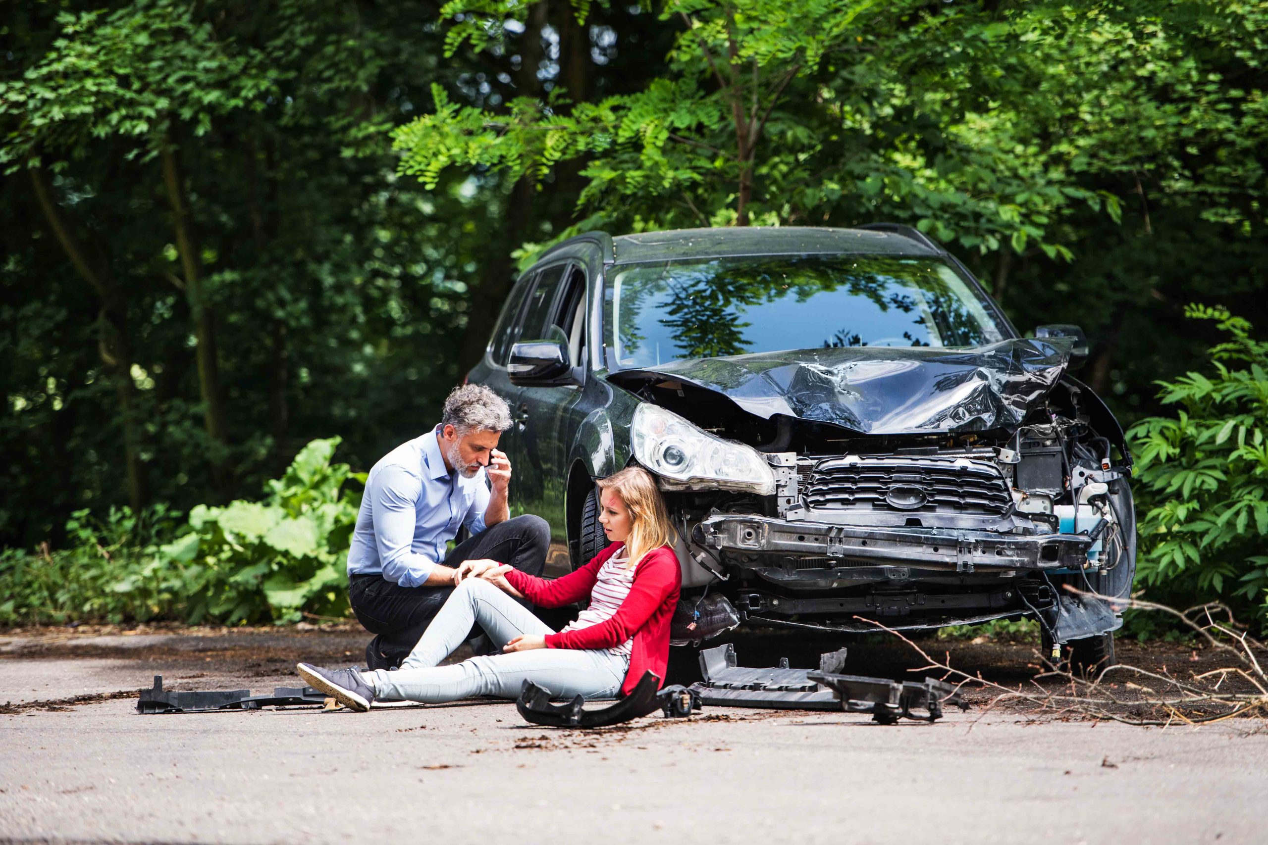 A man kneeling beside a young woman sitting on the ground after a car accident, with a heavily damaged car in the background.