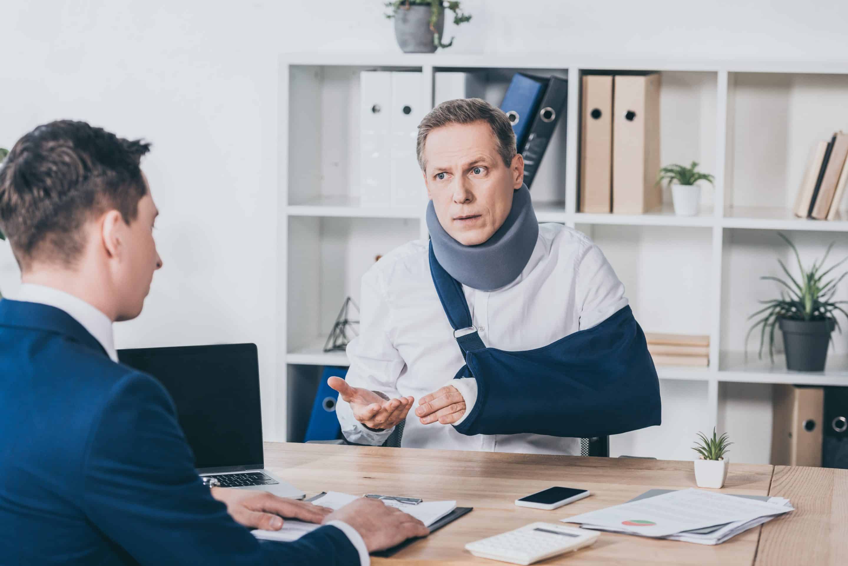 Middle-aged man with a neck brace and arm sling speaking with an attorney in an office, representing legal consultation for personal injury claims