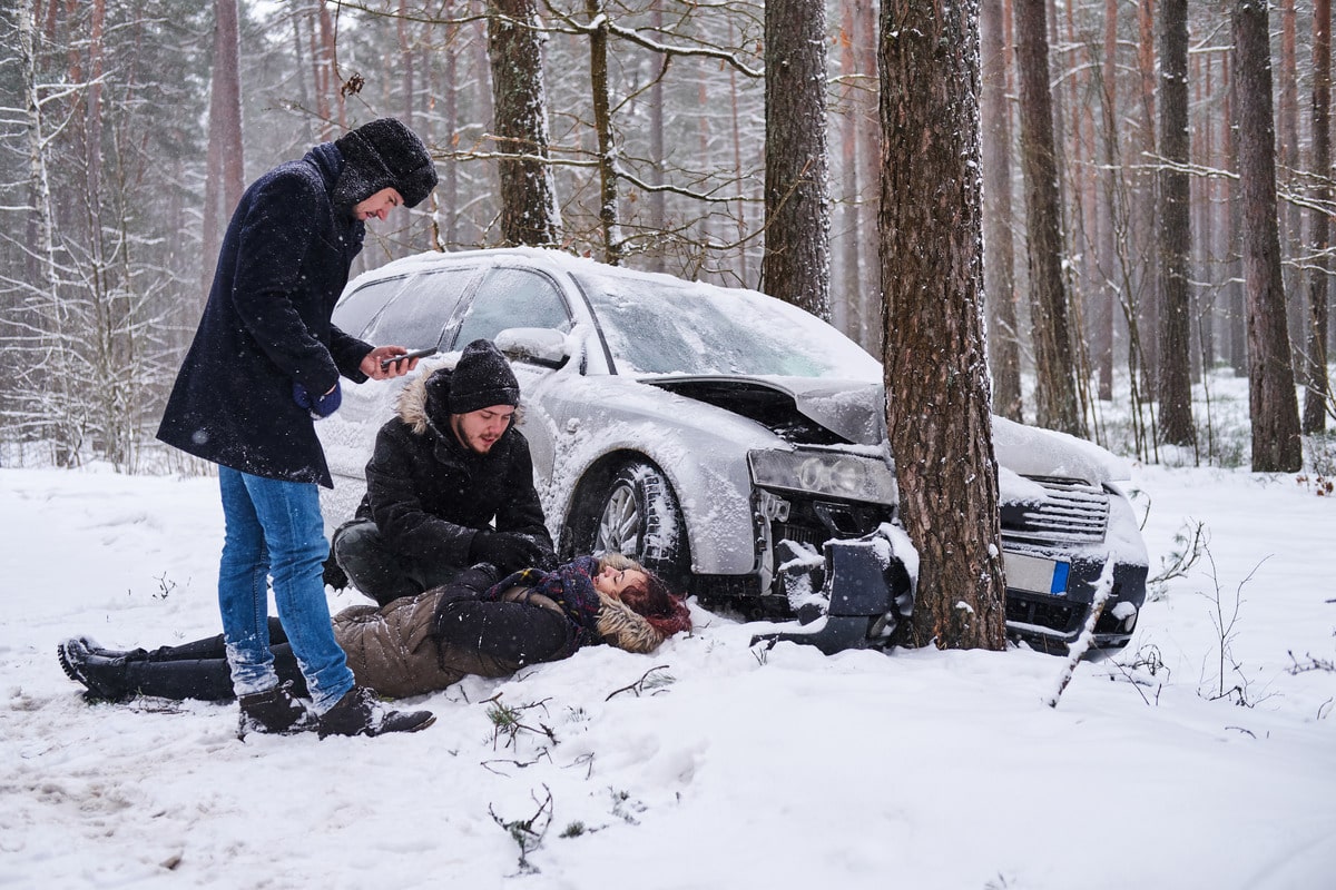 A woman lies on the snowy ground beside a car, indicating a possible accident due to poor road conditions