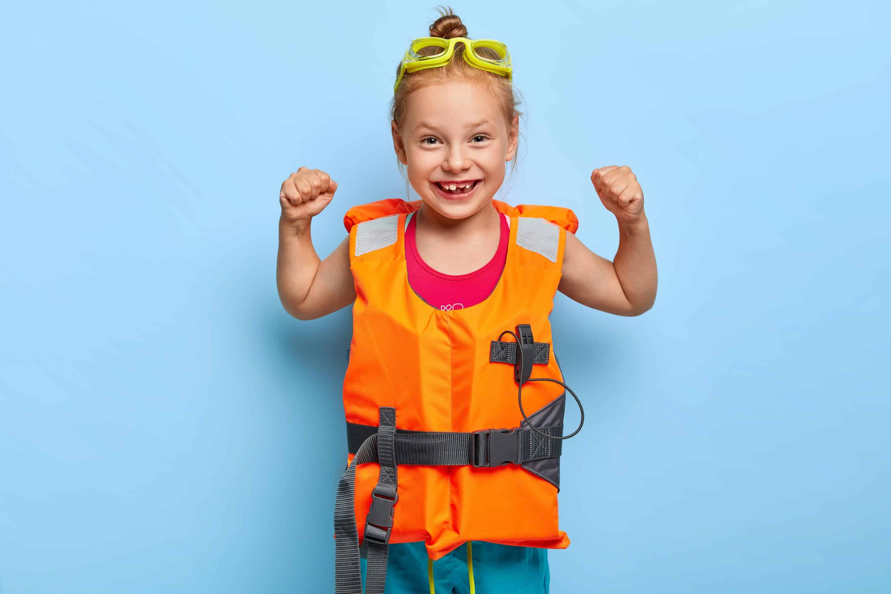 Smiling young girl wearing a bright orange life jacket or personal flotation device and yellow swimming goggles on her head