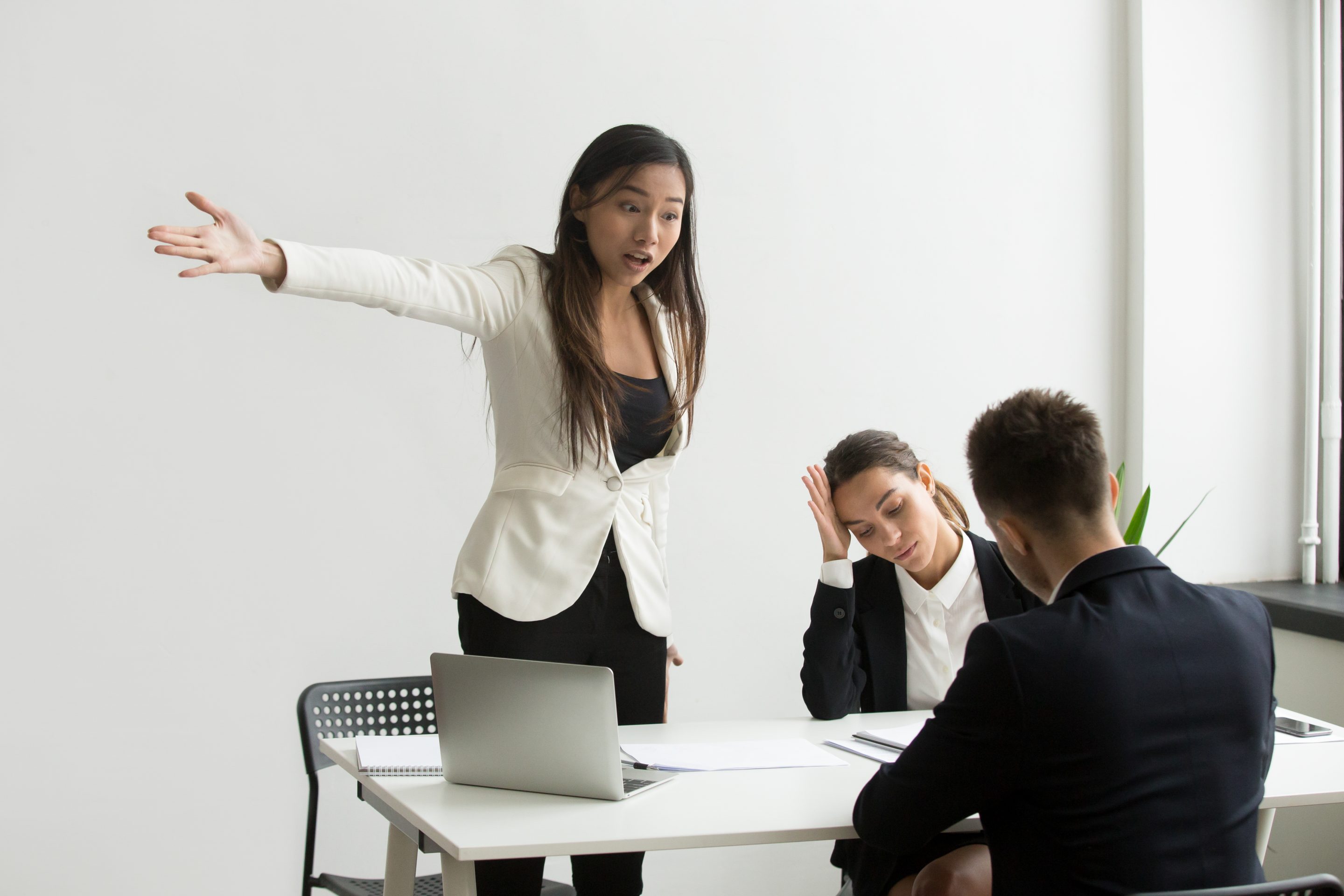 Angry female manager yelling and gesturing to two stressed-looking employees during a tense meeting in a stark white office
