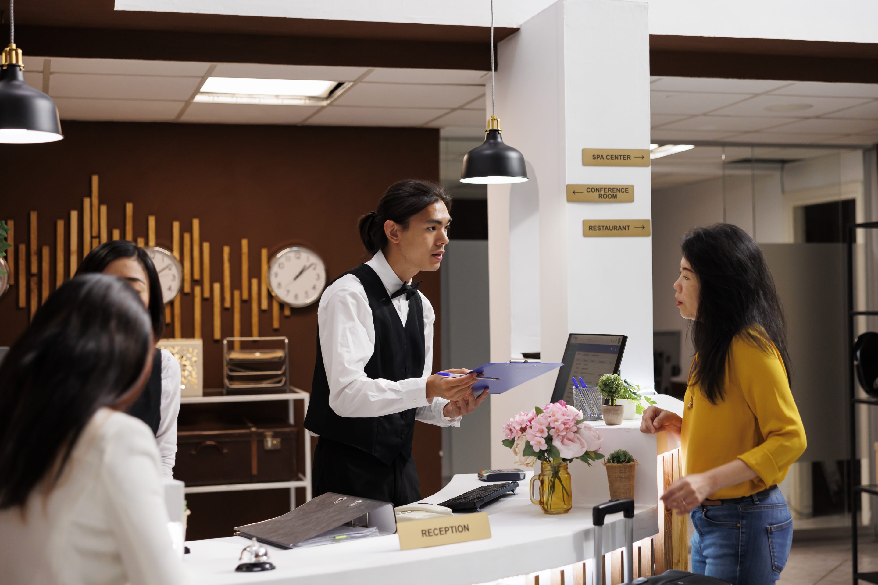 A male hotel receptionist wearing a black vest and white shirt holds a clipboard while speaking with a female guest at a modern white reception desk